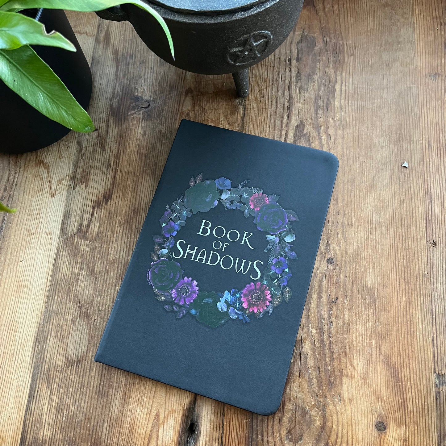 Black notebook titled 'Book of Shadows' with floral design on a wooden surface