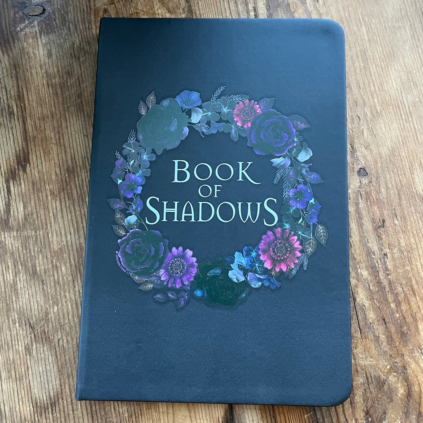 Dark blue notebook with floral design and 'Book of Shadows' text on a wooden surface