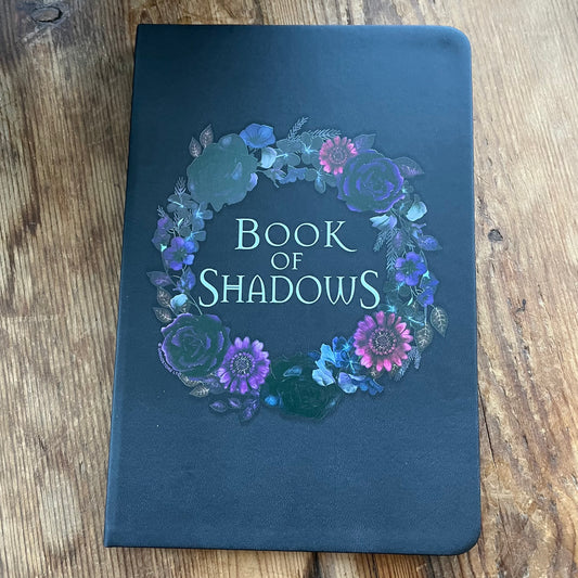 Dark blue notebook with floral design and 'Book of Shadows' text on a wooden surface