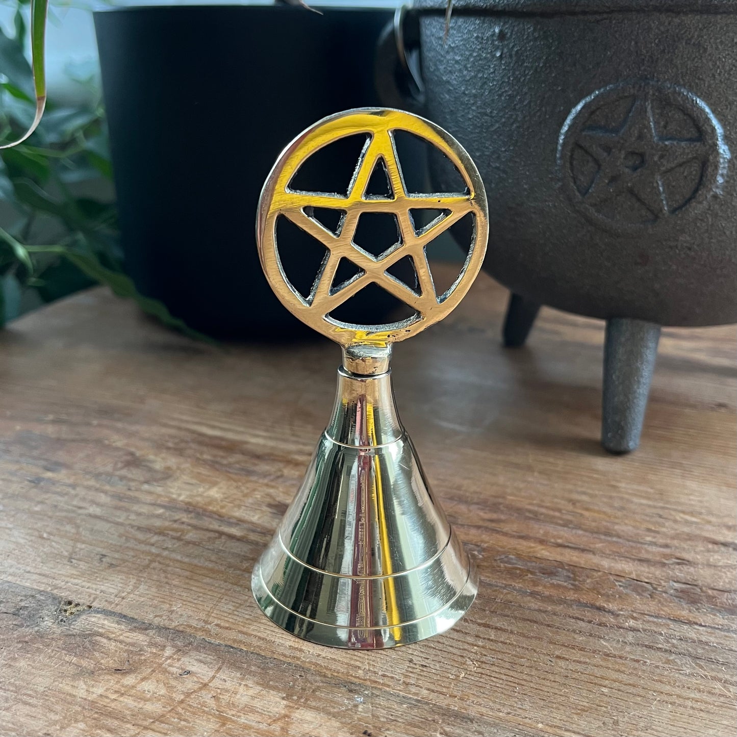 Brass bell with pentagram design on a wooden surface with a black cauldron in the background.
