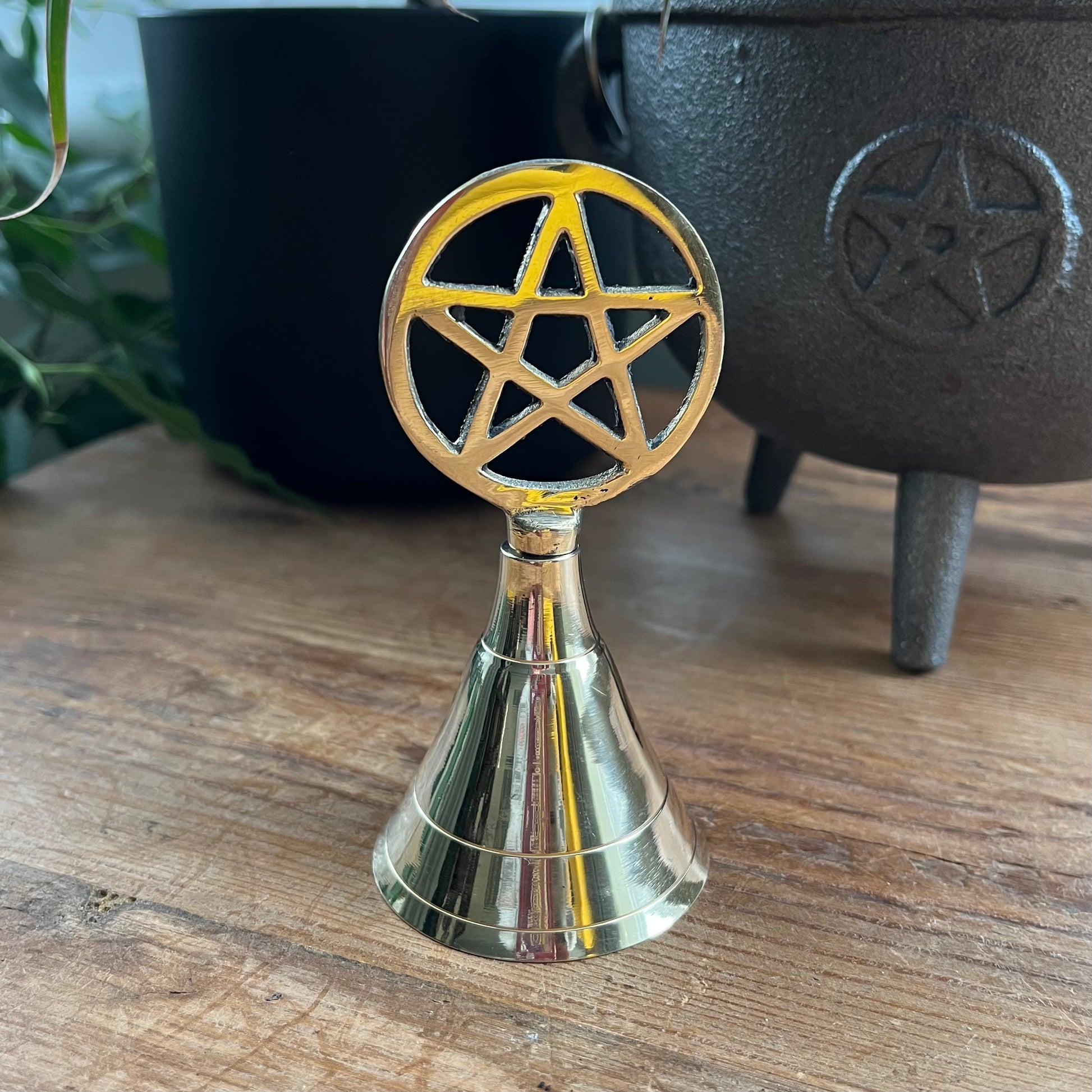 Brass bell with pentagram design on a wooden surface with a black cauldron in the background.