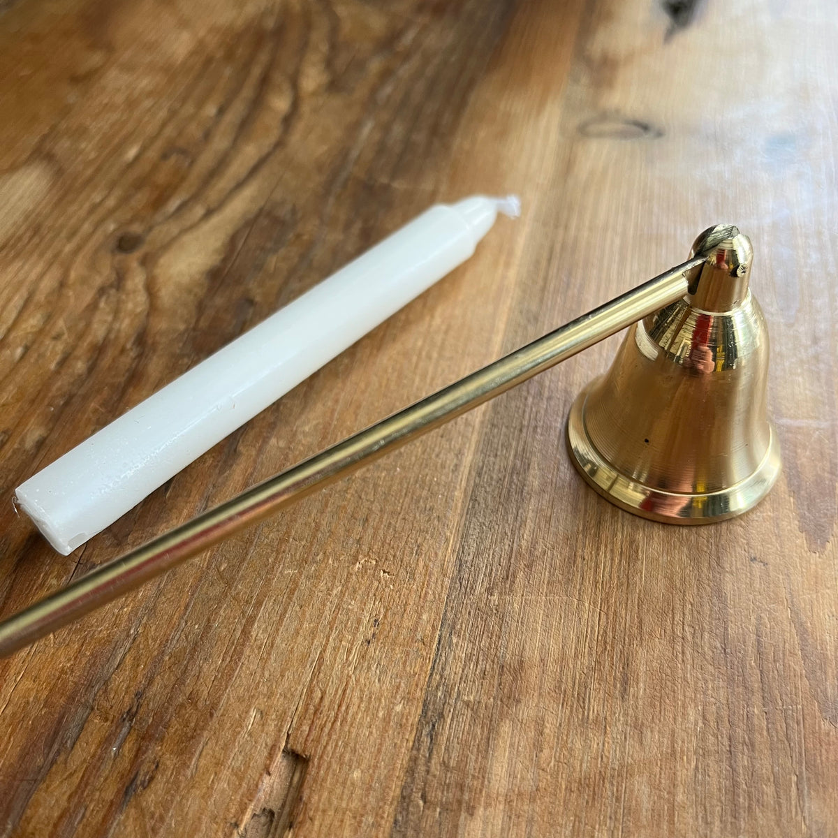 A brass candle snuffer with a long metal arm resting on a wooden table next to a white taper candle.