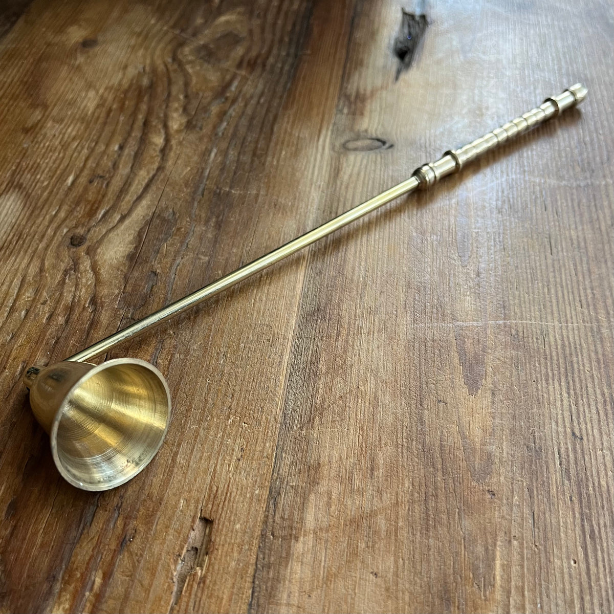 A brass candle snuffer with a long, slender handle and a flared bowl, resting diagonally on a rustic wooden tabletop.