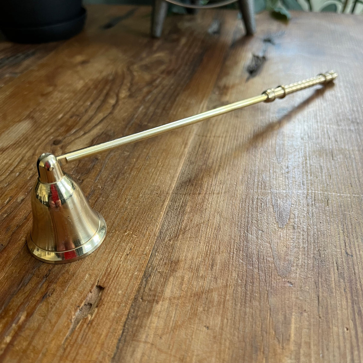 A brass candle snuffer with a bell-shaped head and a long handle, resting on a rustic wooden tabletop.