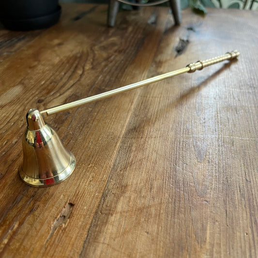 A brass candle snuffer with a bell-shaped head and a long handle, resting on a rustic wooden tabletop.