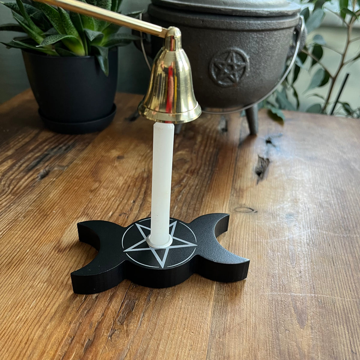 A black wooden triple moon and pentagram candle holder with a brass candle snuffer and white candle, set on a rustic wooden table with potted plants in the background.