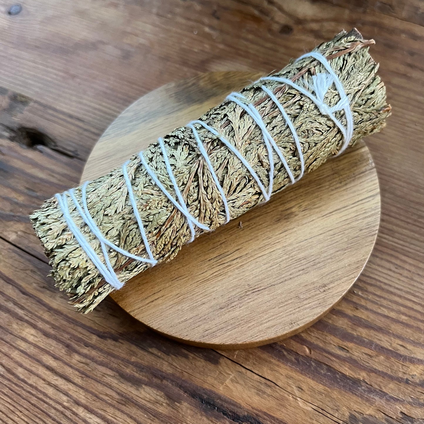 A dried herb smudge bundle wrapped in white string, resting diagonally on a round wooden coaster over a rustic dark wood tabletop.