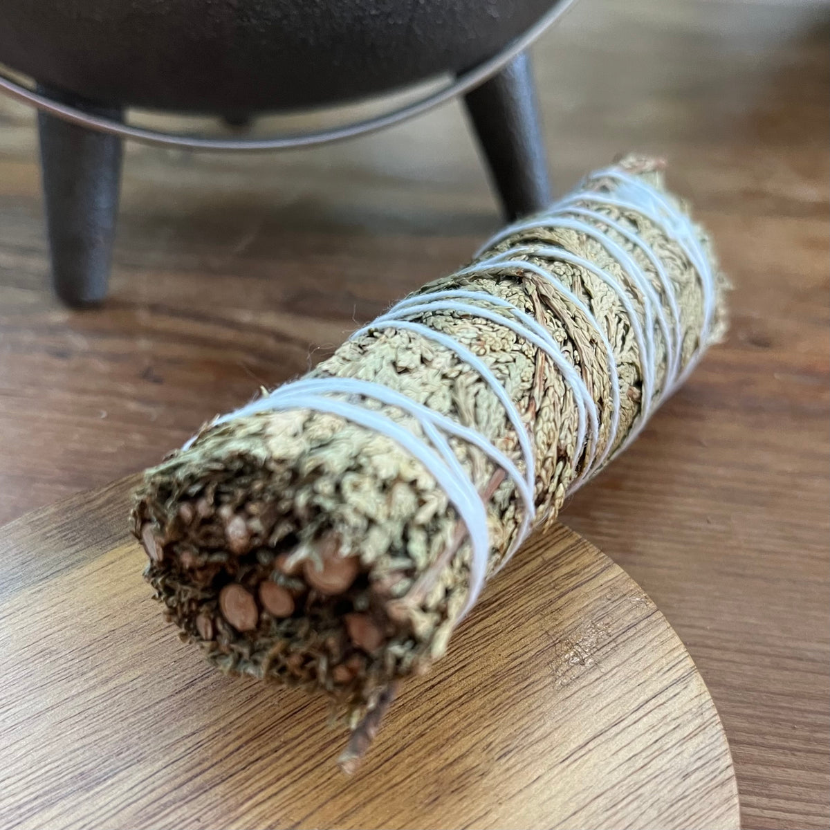 A dried herb smudge bundle wrapped in white string, resting on a wooden surface with a cast iron pot blurred in the background.