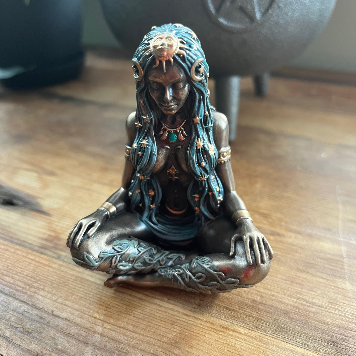 A bronze-finished figurine of a seated goddess with long blue hair, turquoise and gold jewelry, and intricate tattoos, displayed on a wooden tabletop.