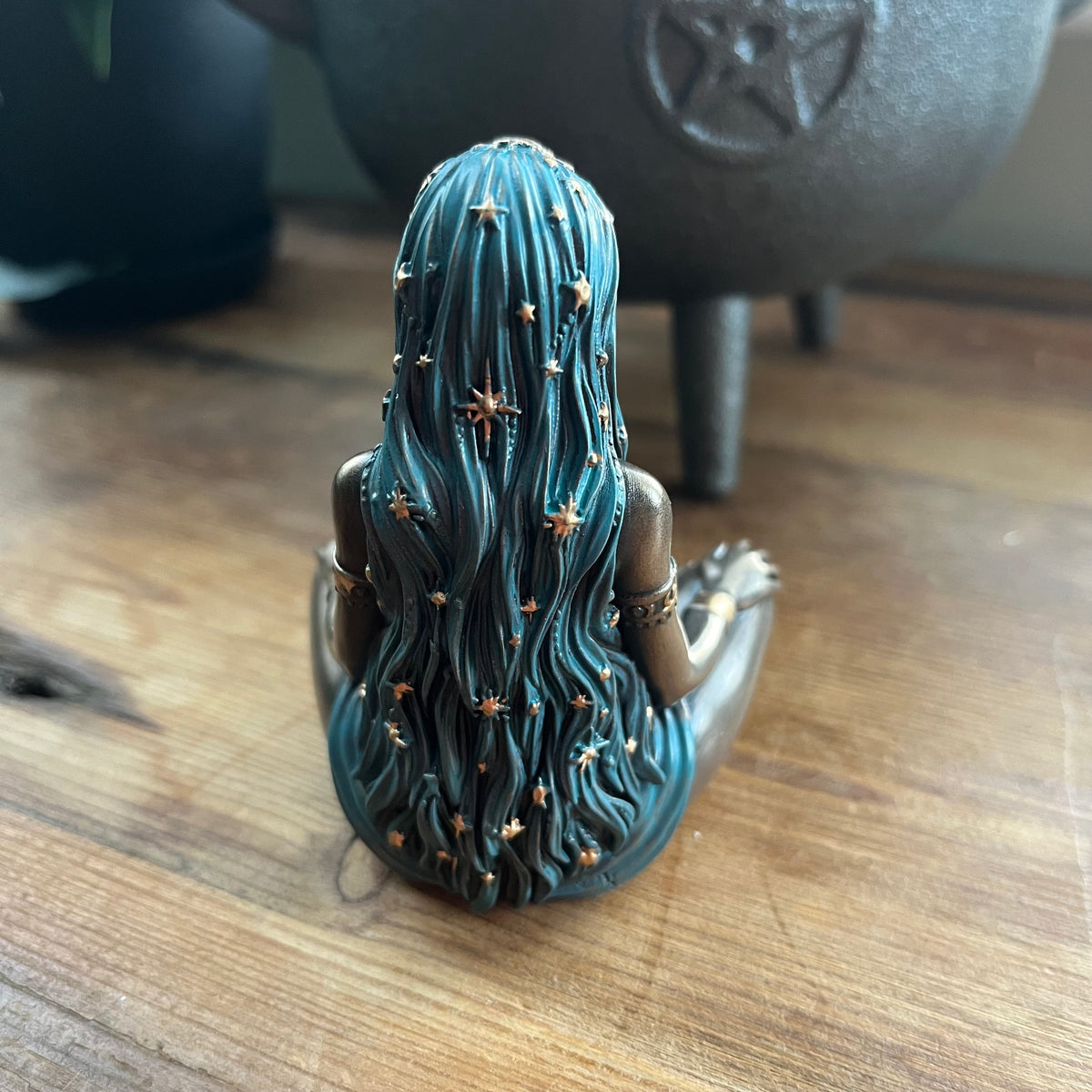 A small figurine of a seated female figure with long teal-blue hair adorned with stars, shown from the back on a wooden tabletop with a dark metal cauldron in the blurred background.