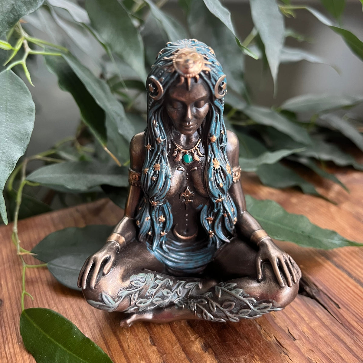 A bronze-finished figurine of a meditating female deity with turquoise blue hair and small gold accents, displayed on a wooden surface surrounded by green leaves.