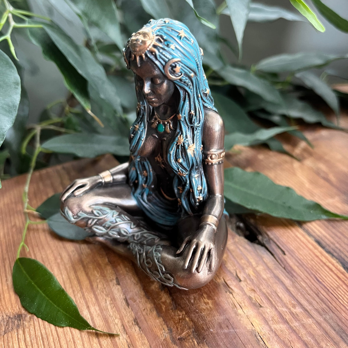 A bronze-finished figurine of a meditating female deity with turquoise blue hair and gold celestial details, displayed on a rustic wooden surface surrounded by green leaves.