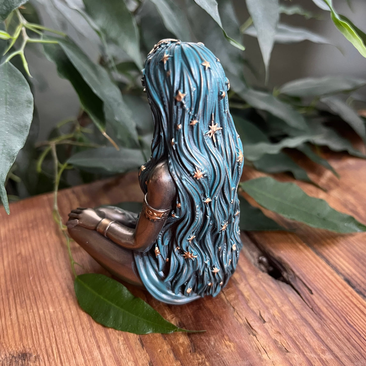 A decorative figurine of a seated woman with long wavy teal-blue hair adorned with small gold stars, displayed on a wooden surface with green leaves in the background.