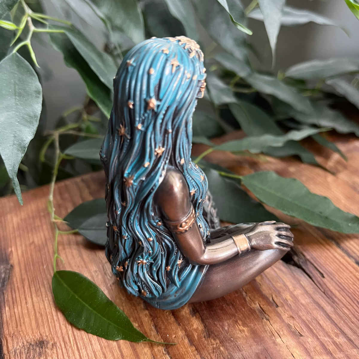 A bronze-finished figurine of a seated woman with long, wavy turquoise-blue hair adorned with small gold stars, displayed on a rustic wooden surface surrounded by green leaves.