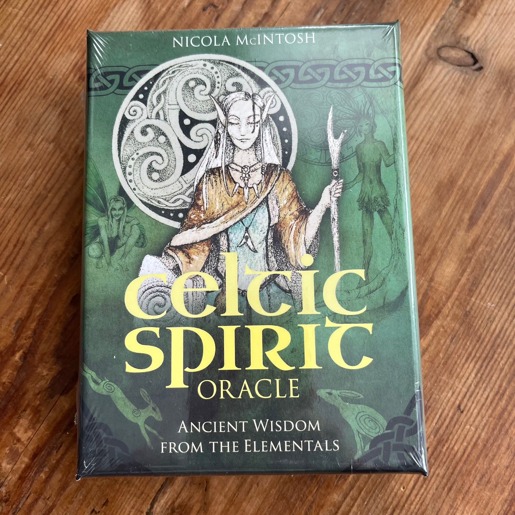 Celtic Spirit Oracle card deck box by Nicola McIntosh, featuring a green cover with an illustration of a Celtic figure and large yellow title text, photographed on a wooden surface.