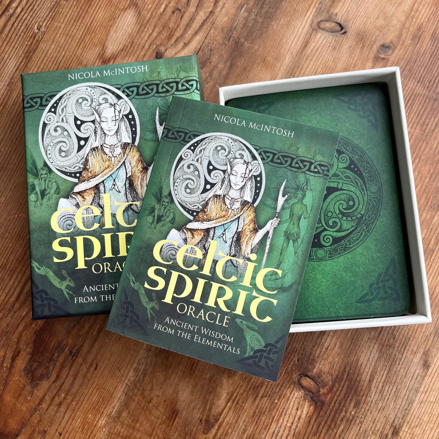 Celtic Spirit Oracle card deck and box set by Nicola McIntosh, featuring green illustrated card backs and boxes on a wooden tabletop.
