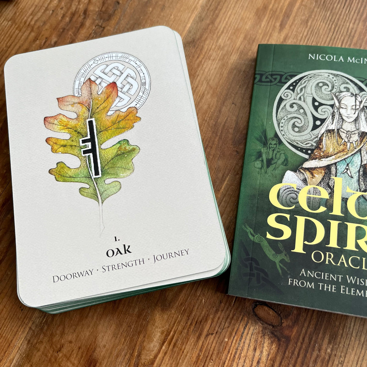 A Celtic Spirit Oracle card deck box sits on a wooden table next to its green book-style book cover, which features an illustration of a woman and the title 'celtic spirit oracles'.