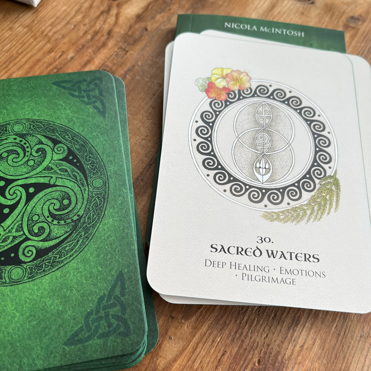A deck of oracle or tarot-style cards on a wooden surface, showing the back of a card with green Celtic knotwork and the front of another card titled "30. SACRED WATERS" with a white background, black line art, and small floral accents.