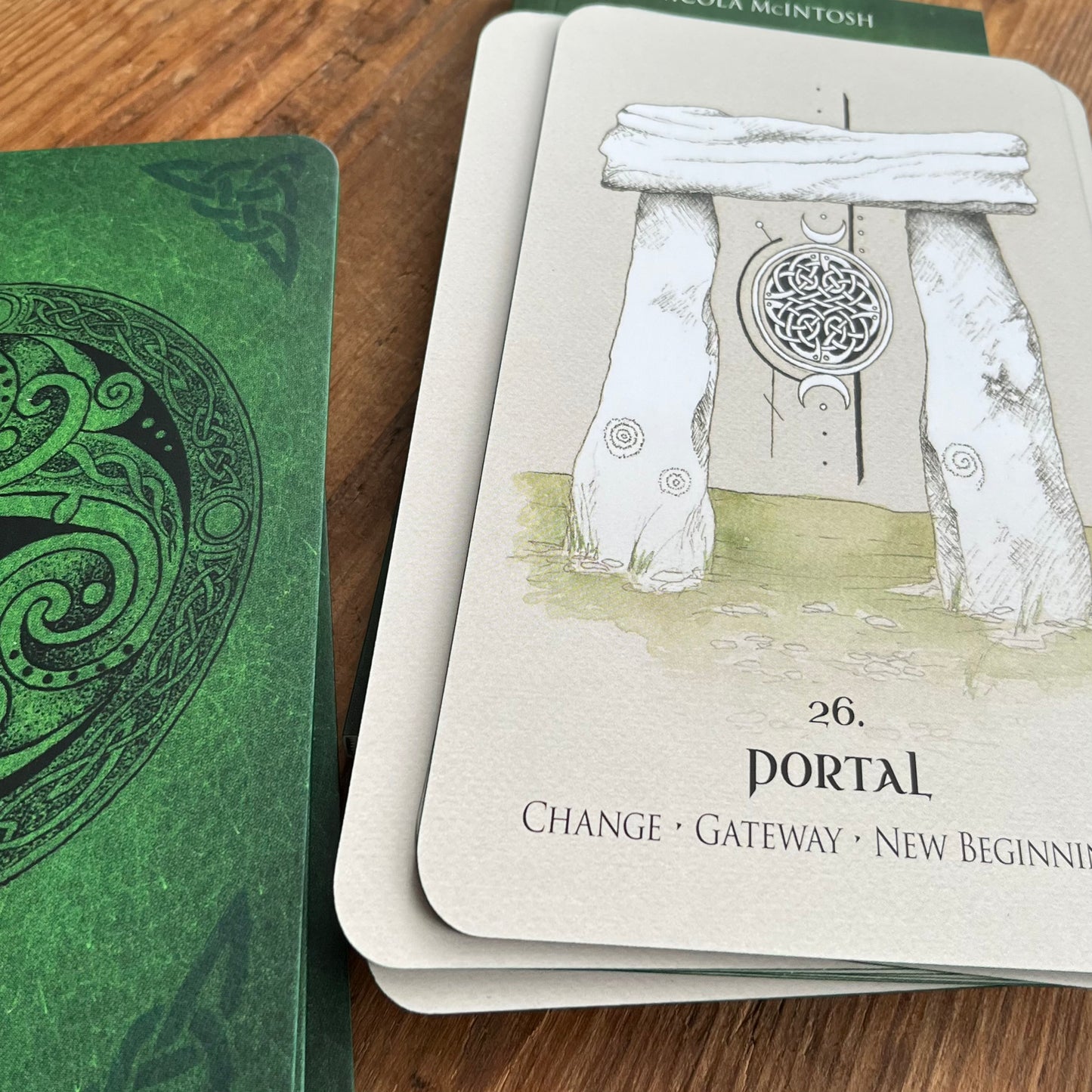 A tarot card deck on a wooden surface, showing the 'Portal' card with an illustration of stone pillars and a circular symbol, alongside another green card with Celtic knotwork.