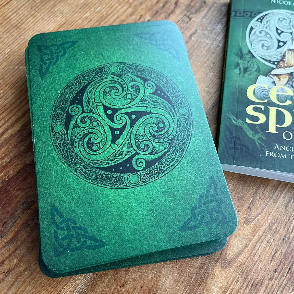 A green Celtic-themed deck of cards with a black triskelion symbol in the center, resting on a wooden table next to its book cover.