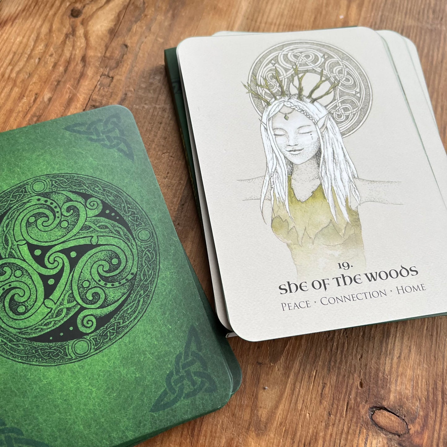 A tarot deck of cards on a wooden table, showing the green illustrated cover of 'The Goddess' card and the white 'She of the Woods' card with Celtic knot details.