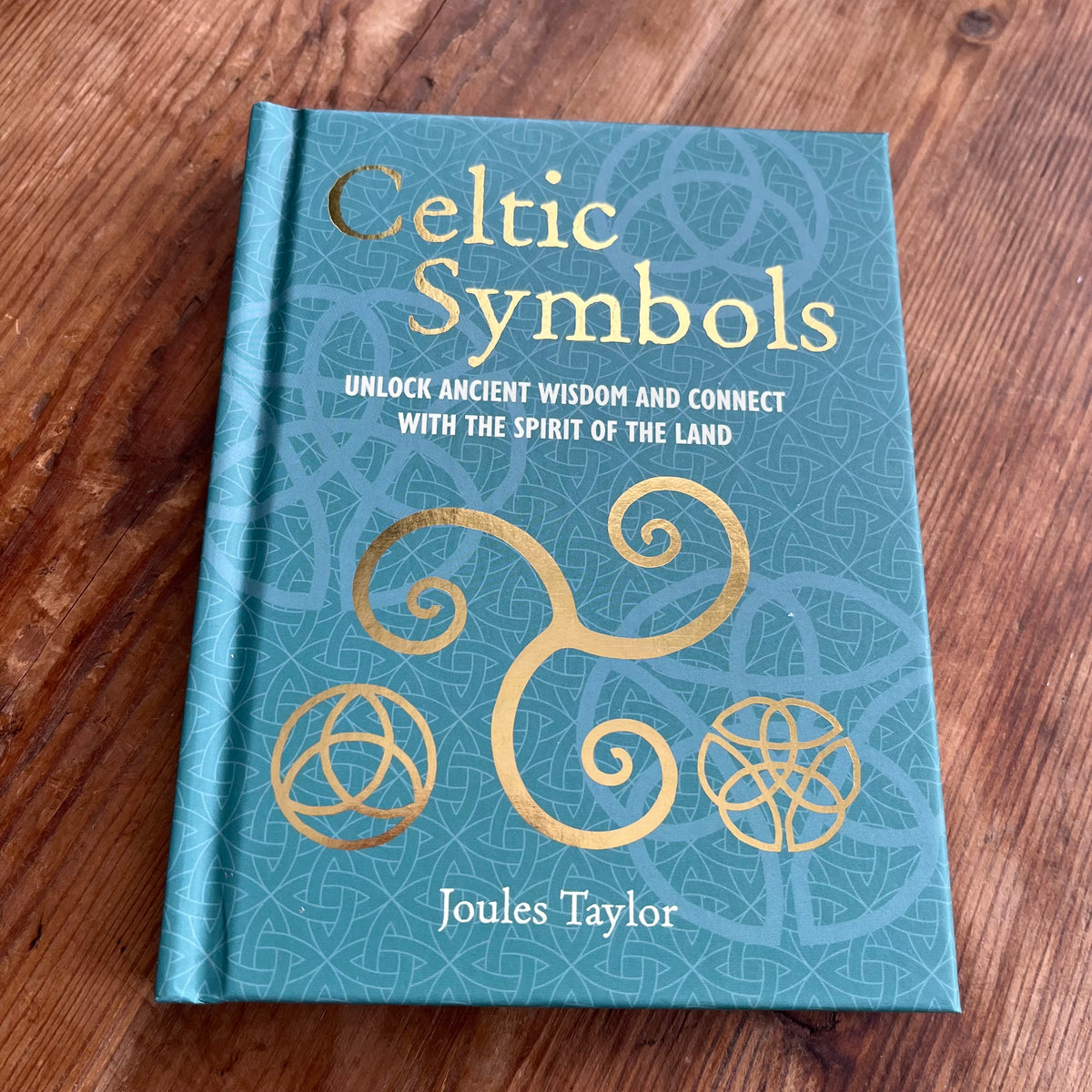 A hardcover book titled 'Celtic Symbols' by Joules Taylor, featuring a teal cover with gold Celtic knot designs and lettering, placed on a wooden surface.