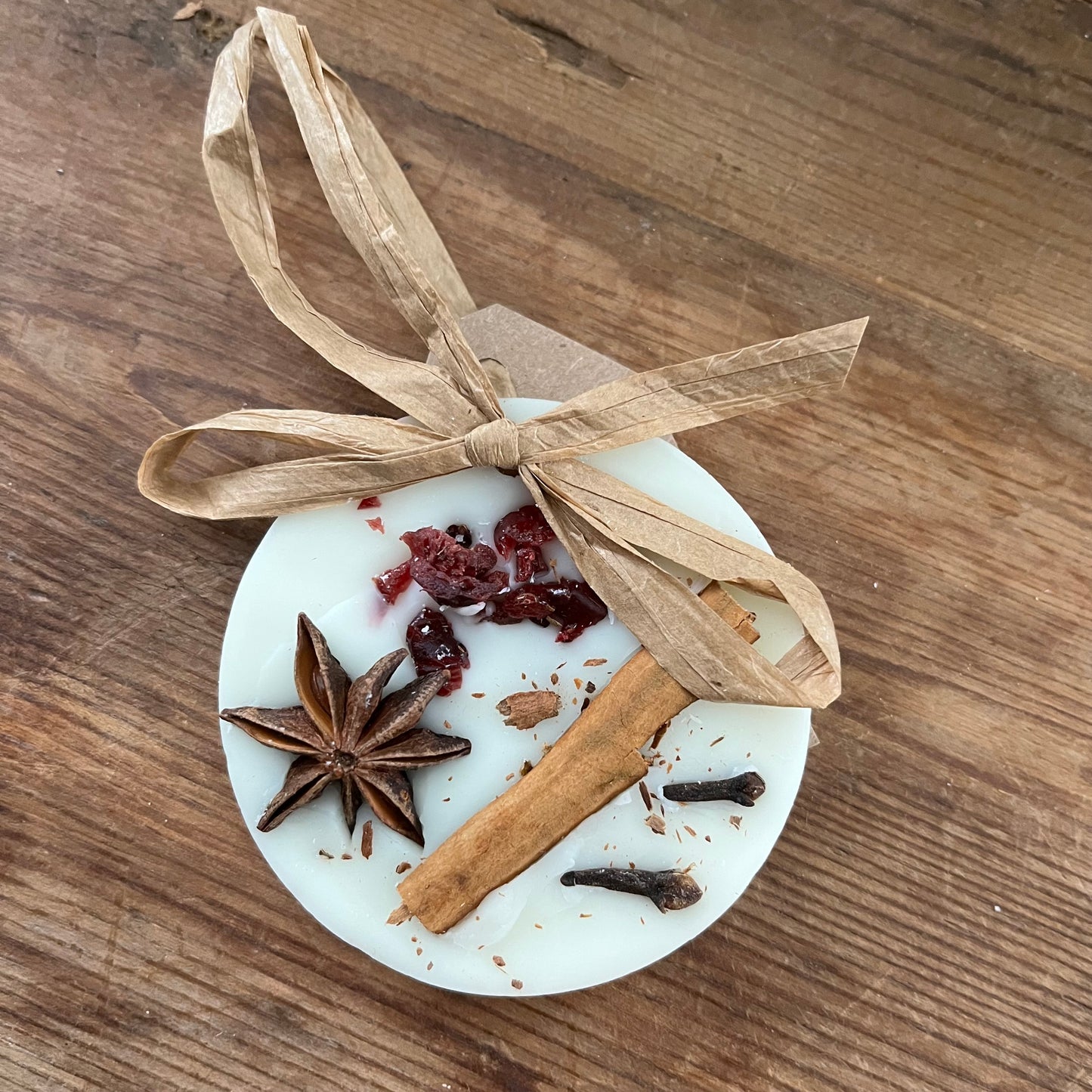 Decorative hanging wax melt item with cinnamon stick, star anise, and cloves on a white base with a brown ribbon on a wooden surface.