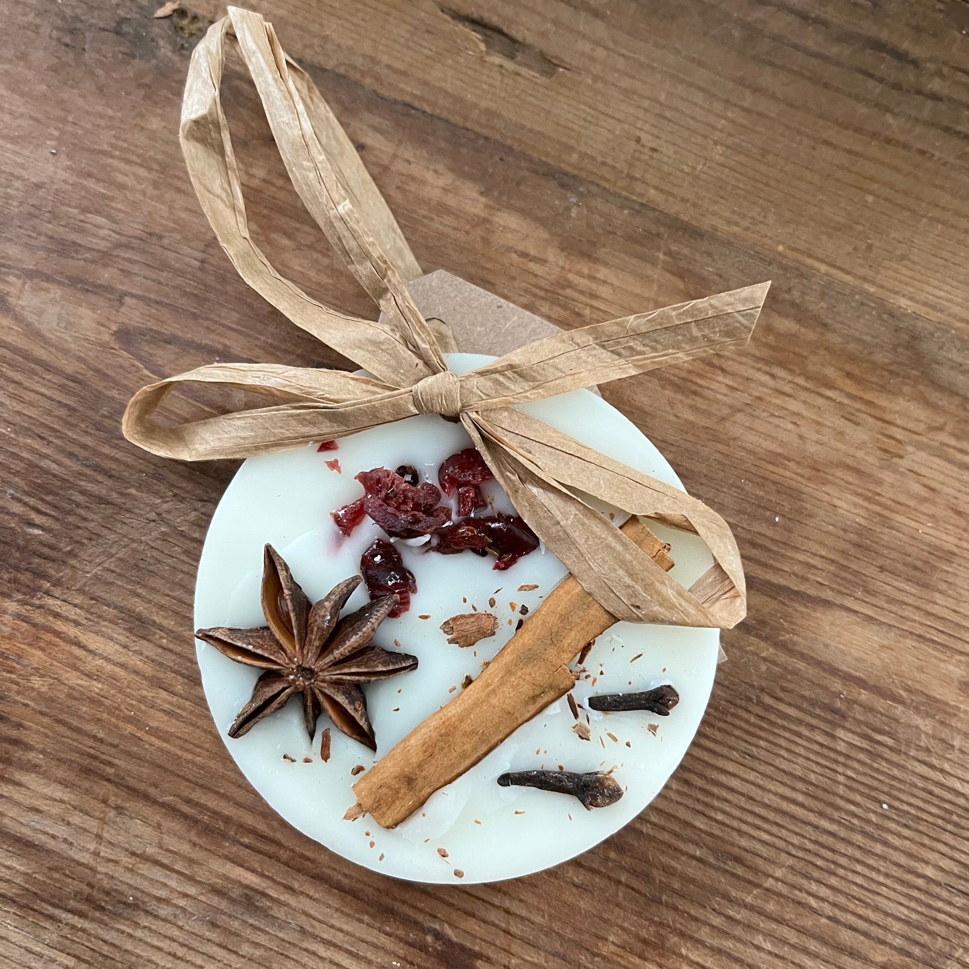 Decorative hanging wax melt item with cinnamon stick, star anise, and cloves on a white base with a brown ribbon on a wooden surface.