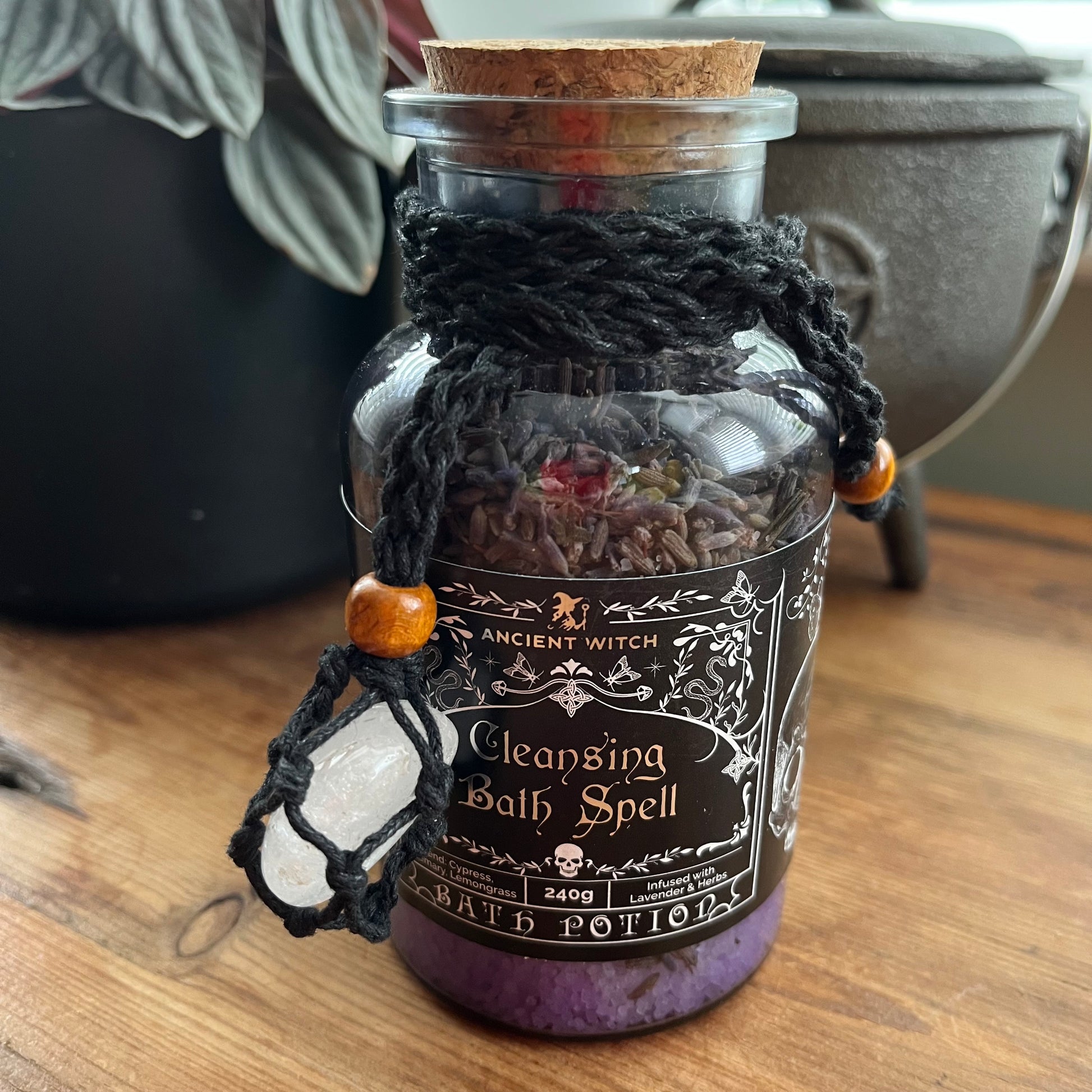 A glass apothecary-style bottle labeled 'Ancient Witch Cleansing Bath Spell' with a cork stopper, black braided cord, wooden beads, and a white crystal pendant, filled with dried herbs and purple powder at the bottom, set on a wooden surface.