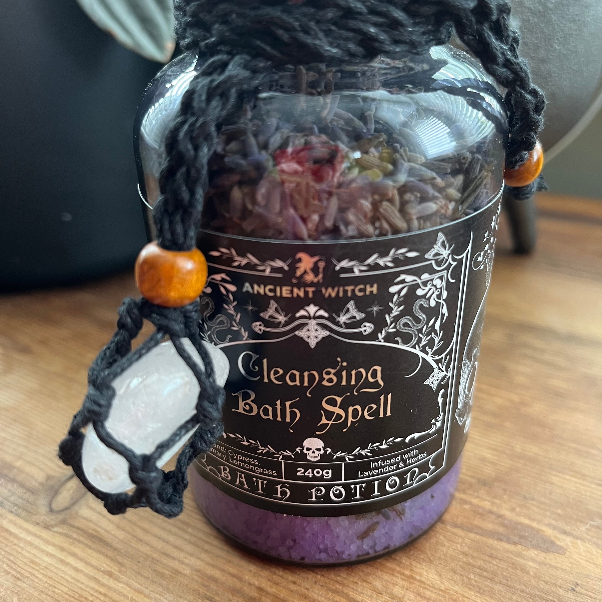 A glass jar labeled 'Ancient Witch Cleansing Bath Spell' filled with purple bath salts and dried botanicals, with a black braided hanging cord, wooden beads, and a white crystal pendant on a wooden surface.