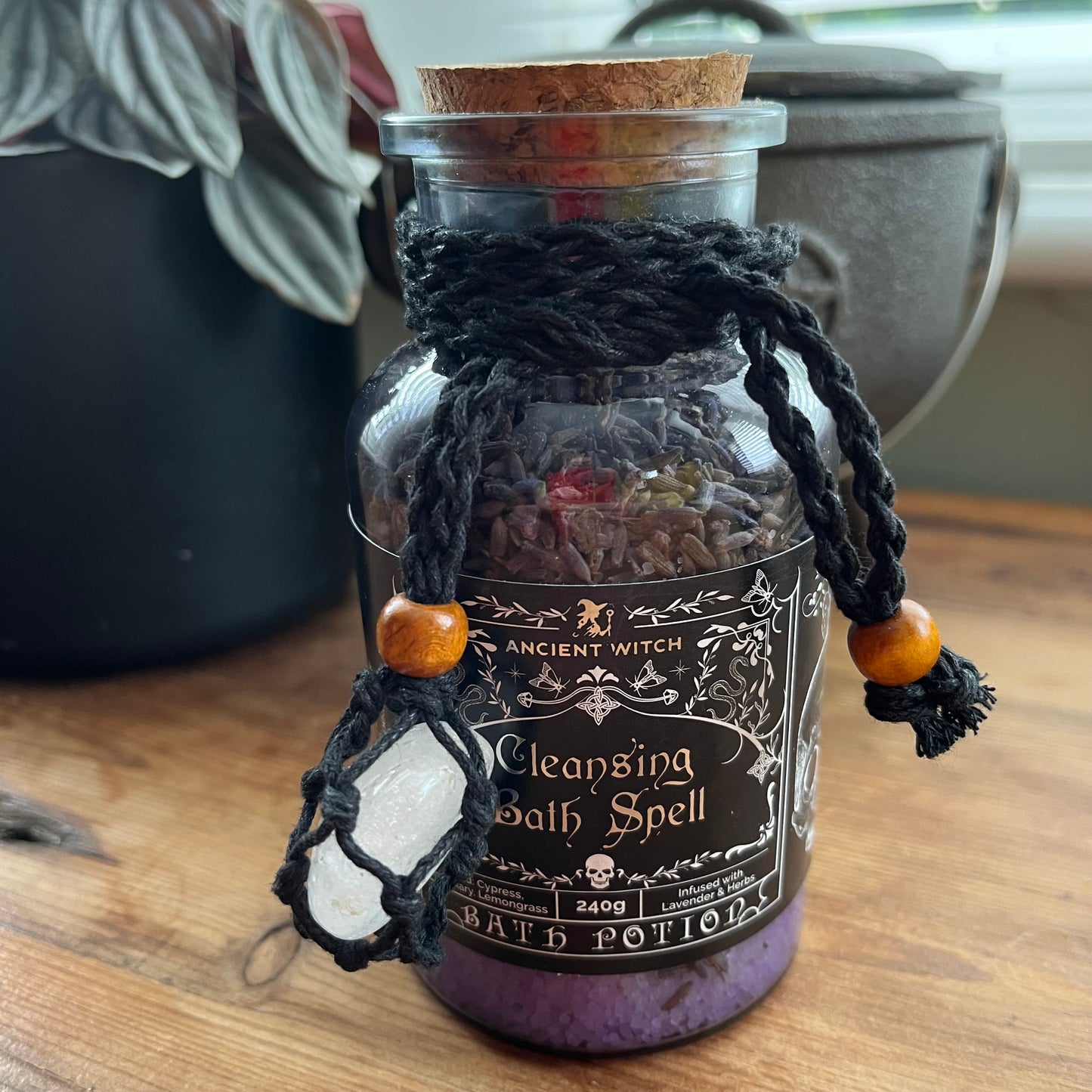 A glass bottle labeled 'Ancient Witch Cleansing Bath Spell' with a cork stopper, black braided cord, wooden beads, and a white crystal pendant, sitting on a wooden surface.
