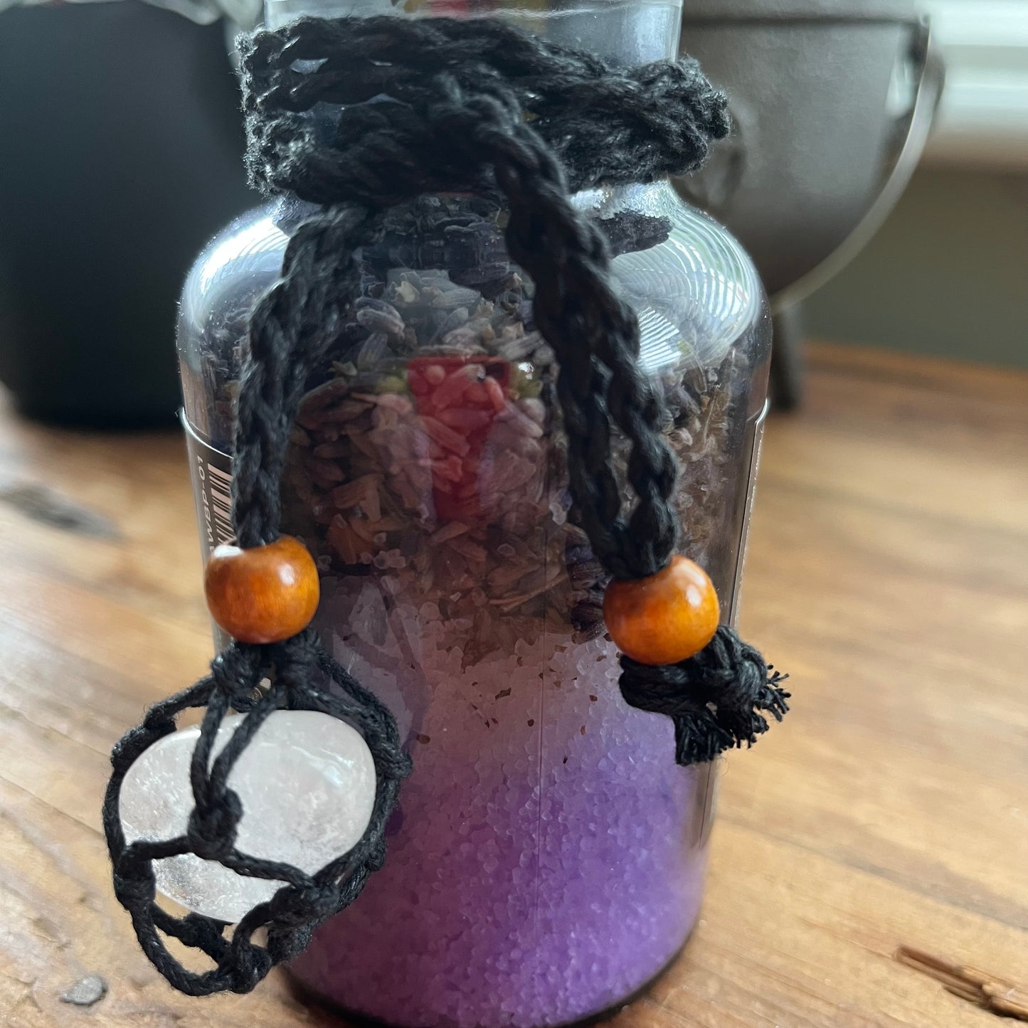 A handmade crystal jar with a purple and lavender layered base, filled with dried herbs, featuring a black macrame cord closure adorned with orange wooden beads and a white quartz. resting on a wooden table.