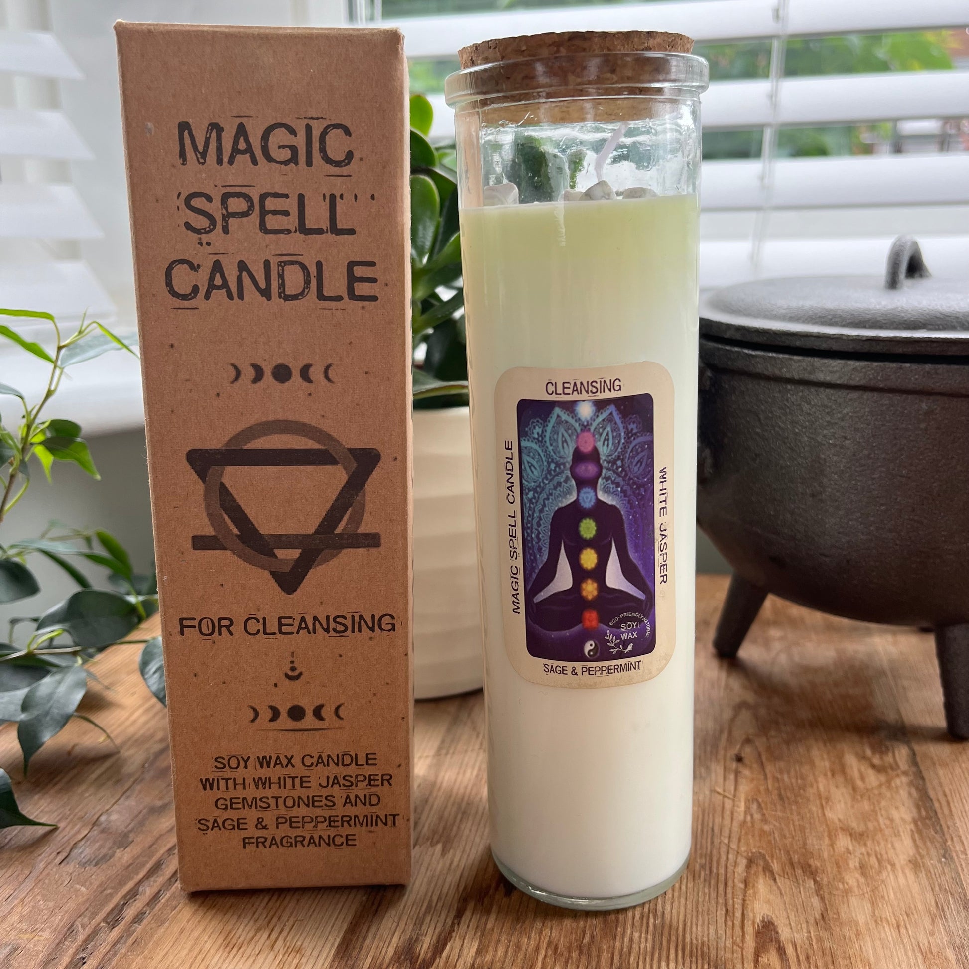 Tall glass spell candle with cork lid. The label on the candle features an image of a seated person and their coloured chakras and the words Magic spell candle, Cleansing, white Jasper and Sage and Peppermint. The candle itself is white with white Jasper gemstones embedded within it. Alongside the glass Magic spell candle is it's brown cardboard box which has the words magic spell candle for Cleansing, soy wax candle with white Jasper gemstones and sage and peppermint fragrance.