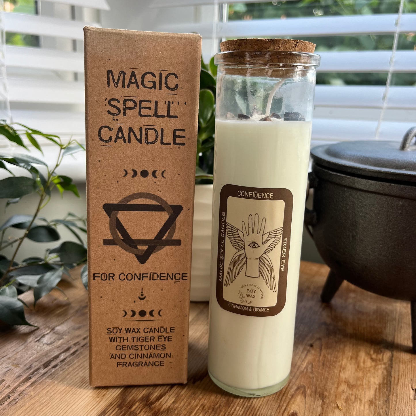 Tall glass spell candle with cork lid. The label on the candle features a winged hand with an eye in its palm and the words Confidence, Tigers eye and Cinnamon. The candle itself is white with Tigers eye gemstones embedded within it. Alongside the glass Magic spell candle is it's brown cardboard box which has the words magic spell candle for confidence, soy wax candle with Tigers eye gemstones and Cinnamon fragrance.