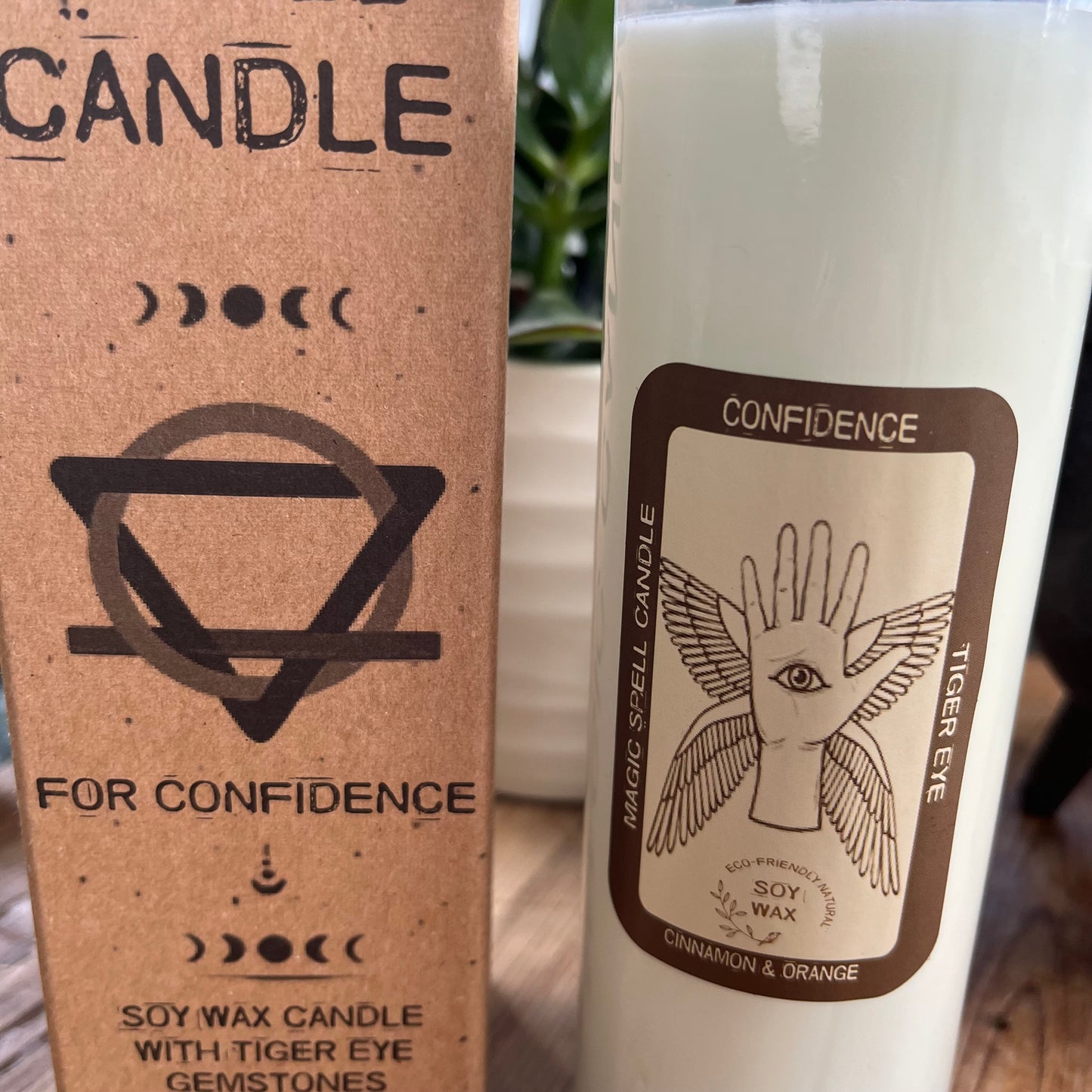 Tall glass spell candle with cork lid. The label on the candle features a winged hand with an eye in its palm and the words Confidence, Tigers eye and Cinnamon. The candle itself is white with Tigers eye gemstones embedded within it. Alongside the glass Magic spell candle is it's brown cardboard box which has the words magic spell candle for confidence, soy wax candle with Tigers eye gemstones and Cinnamon fragrance.