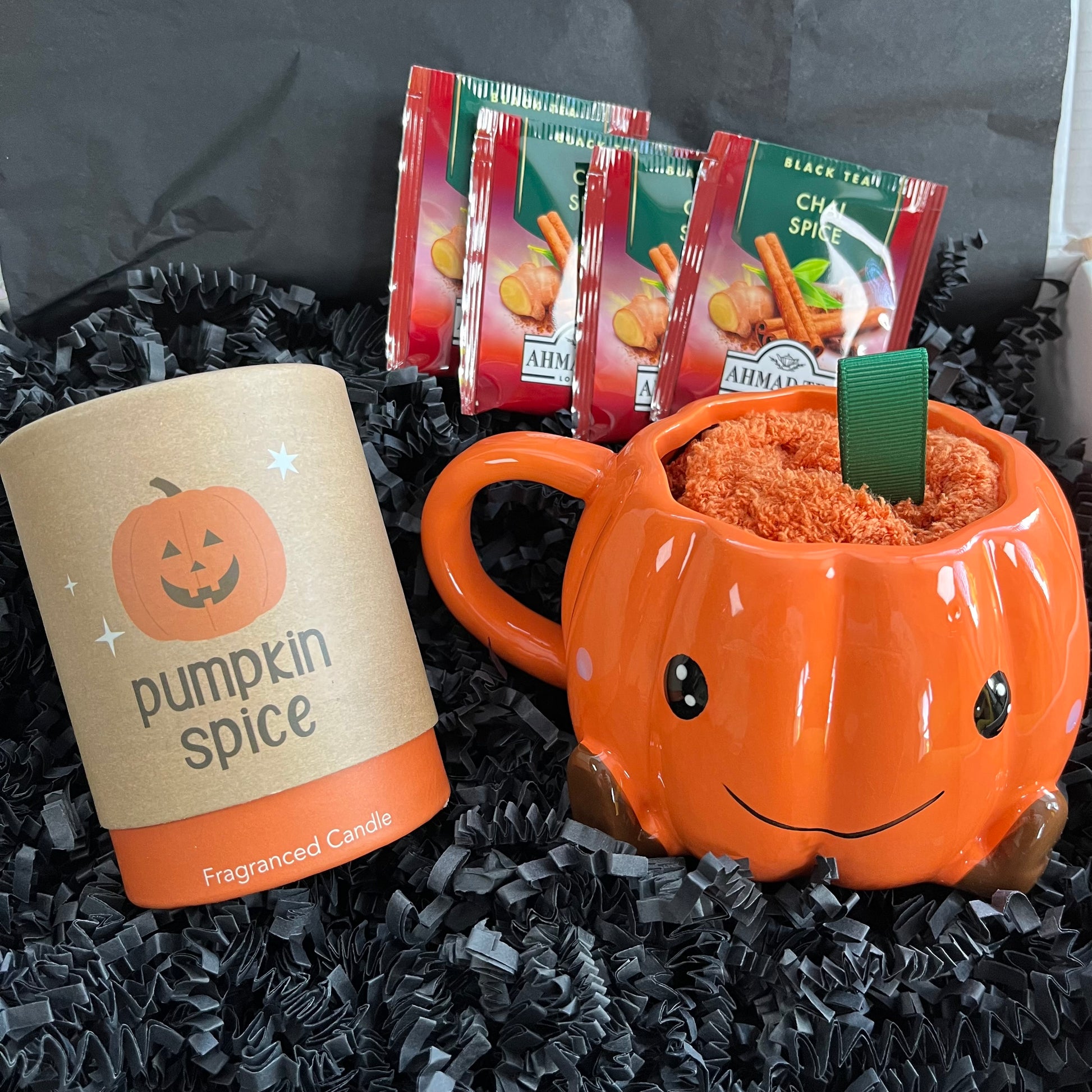 A fall-themed gift set featuring a 'pumpkin spice' scented candle in a kraft paper tube and an orange pumpkin-shaped mug filled with orange cosy socks set in black crinkle paper with AIMAKO black tea packets in the background.