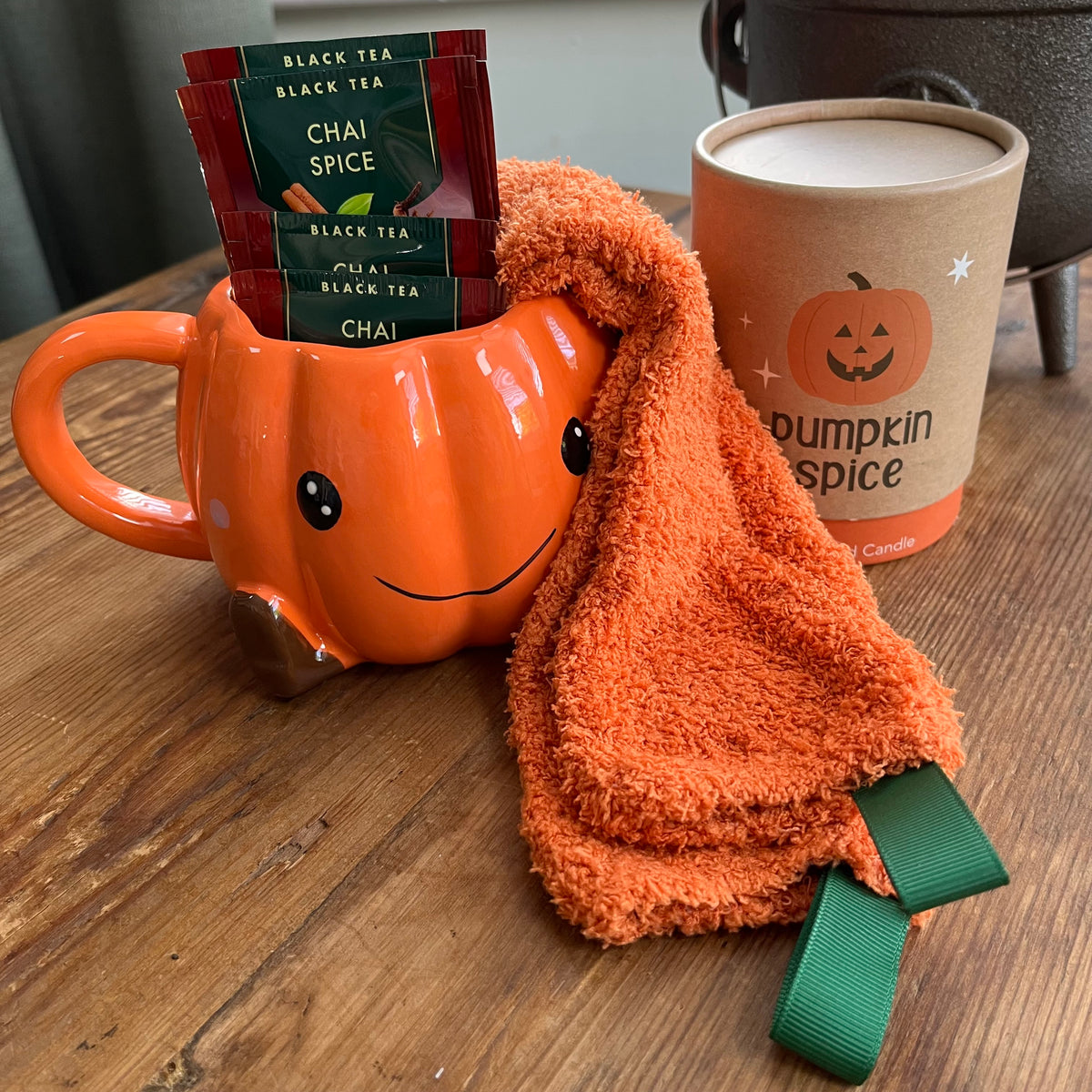 An orange pumpkin-shaped ceramic mug filled with black tea packets, next to an orange cosy socks draped over the mug and a boxed candle labeled pumpkin spice, all arranged on a wooden tabletop.