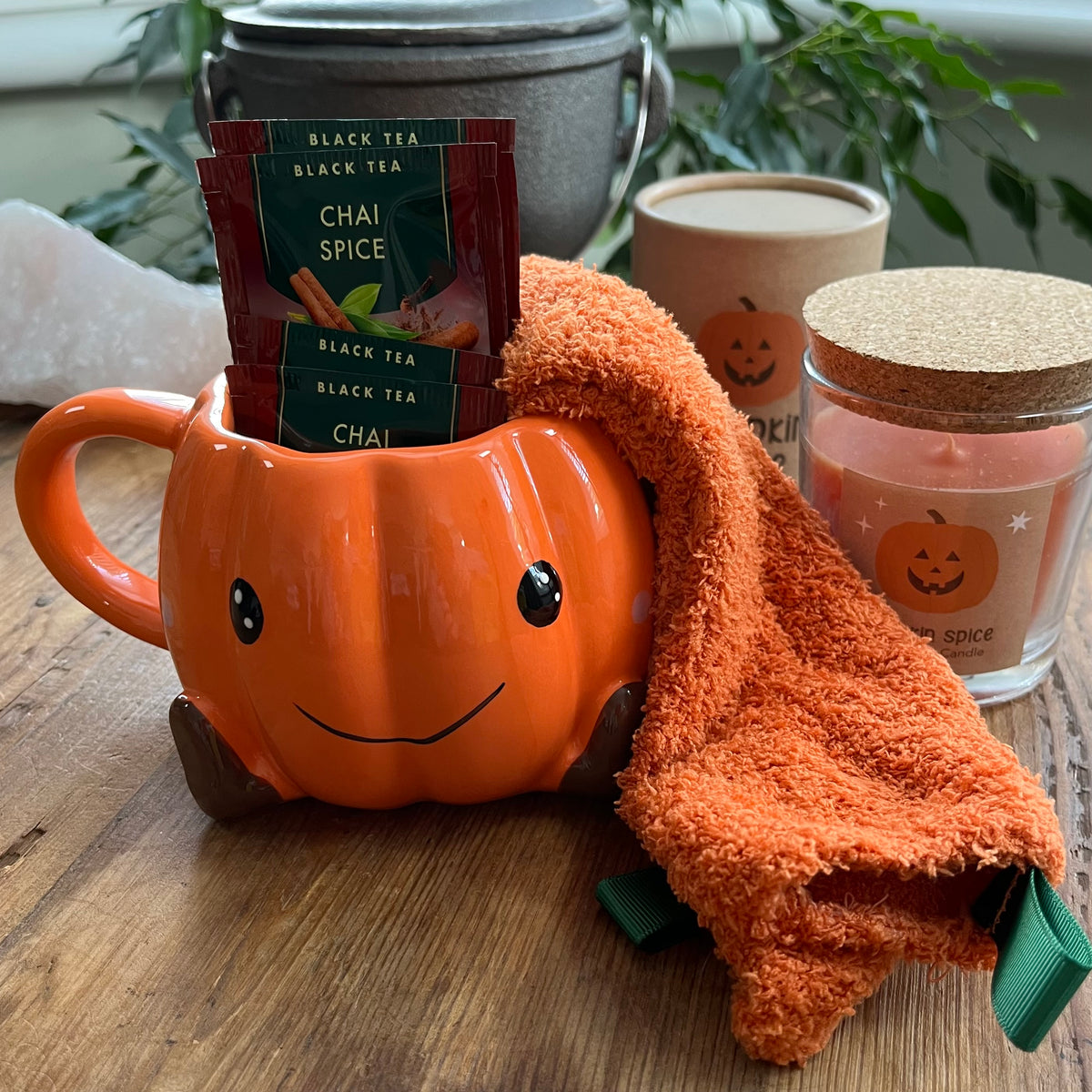 An orange pumpkin-shaped ceramic mug with a smiling face, filled with packets of black tea and draped with an orange cosy pair of socks. The mug sits on a wooden table with candles and plants in the background.