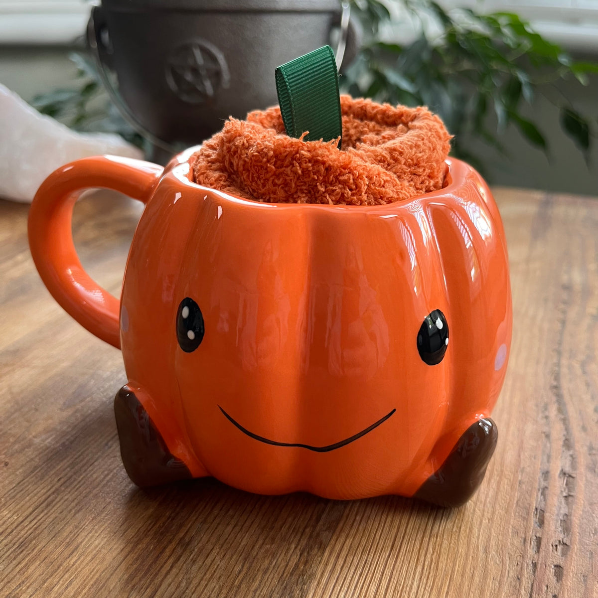 An orange pumpkin-shaped ceramic mug with a smiling face, sitting on a wooden table. Inside the mug, a textured orange socks is tucked into the top opening, with a small green ribbon loop visible.