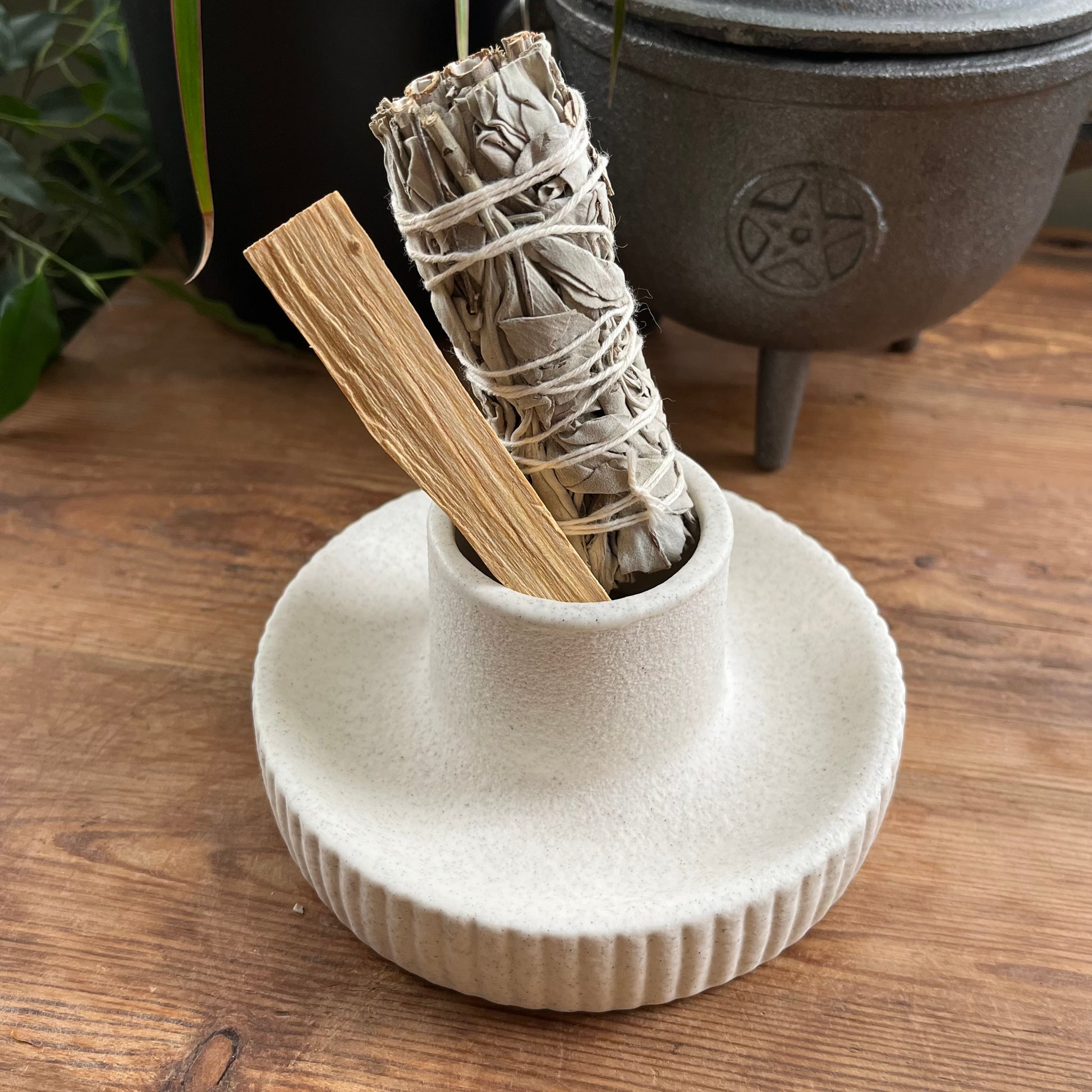White ceramic holder with a smoldering sage stick and Palo Santo stick on a wooden surface.