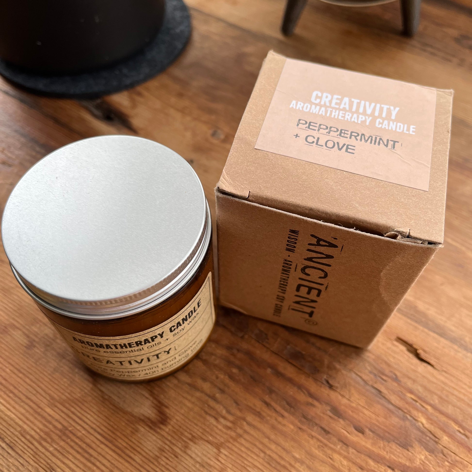 A brown glass jar candle with a silver lid sits next to its kraft paper box on a wooden table. The packaging is labeled 'ANCIENT' and 'CREATIVITY AROMA THERAPY CANDLE' with scent details 'PEPPERMINT + CLOVE'.