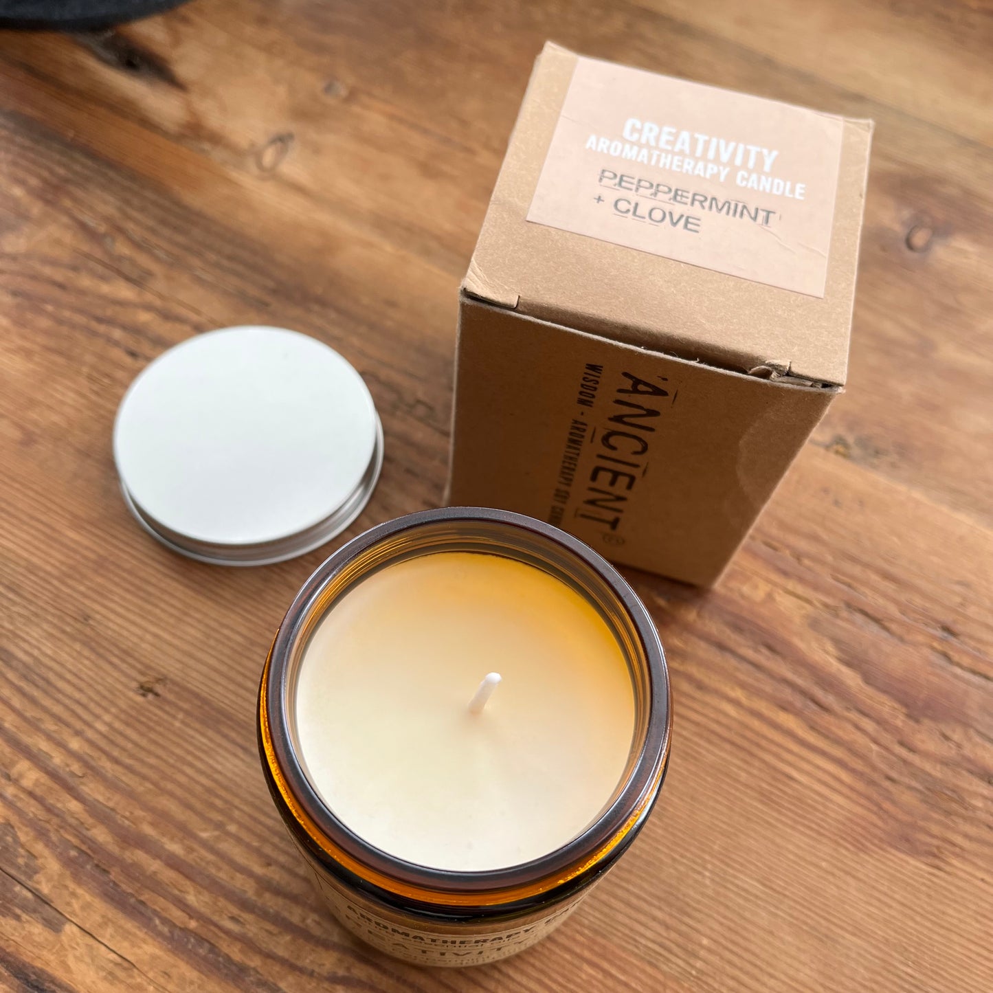 An open amber glass jar candle with cream wax sits on a wooden surface next to its cardboard box and a white lid. The box reads 'ANCIENT' and 'CREATIVITY AROMATHERAPY CANDLE' with 'PEPPERMINT + CLOVE' visible on the top label.