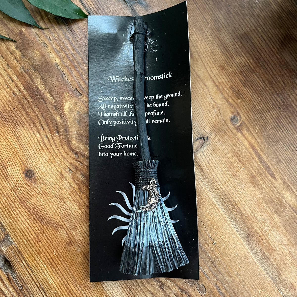 A black witch's decorative broomstick with a silver crescent moon charm, displayed on its printed black backing card on a wooden tabletop.