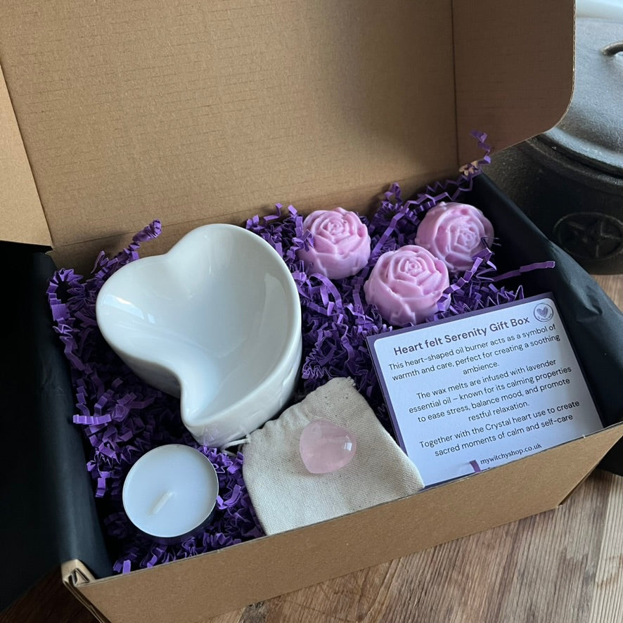 White ceramic heart shaped oil burner, a white tealight candle, 3 pink flower shaped wax melts and a small pink rose quartz heart are presented in a gift box alongside 2 information cards. 