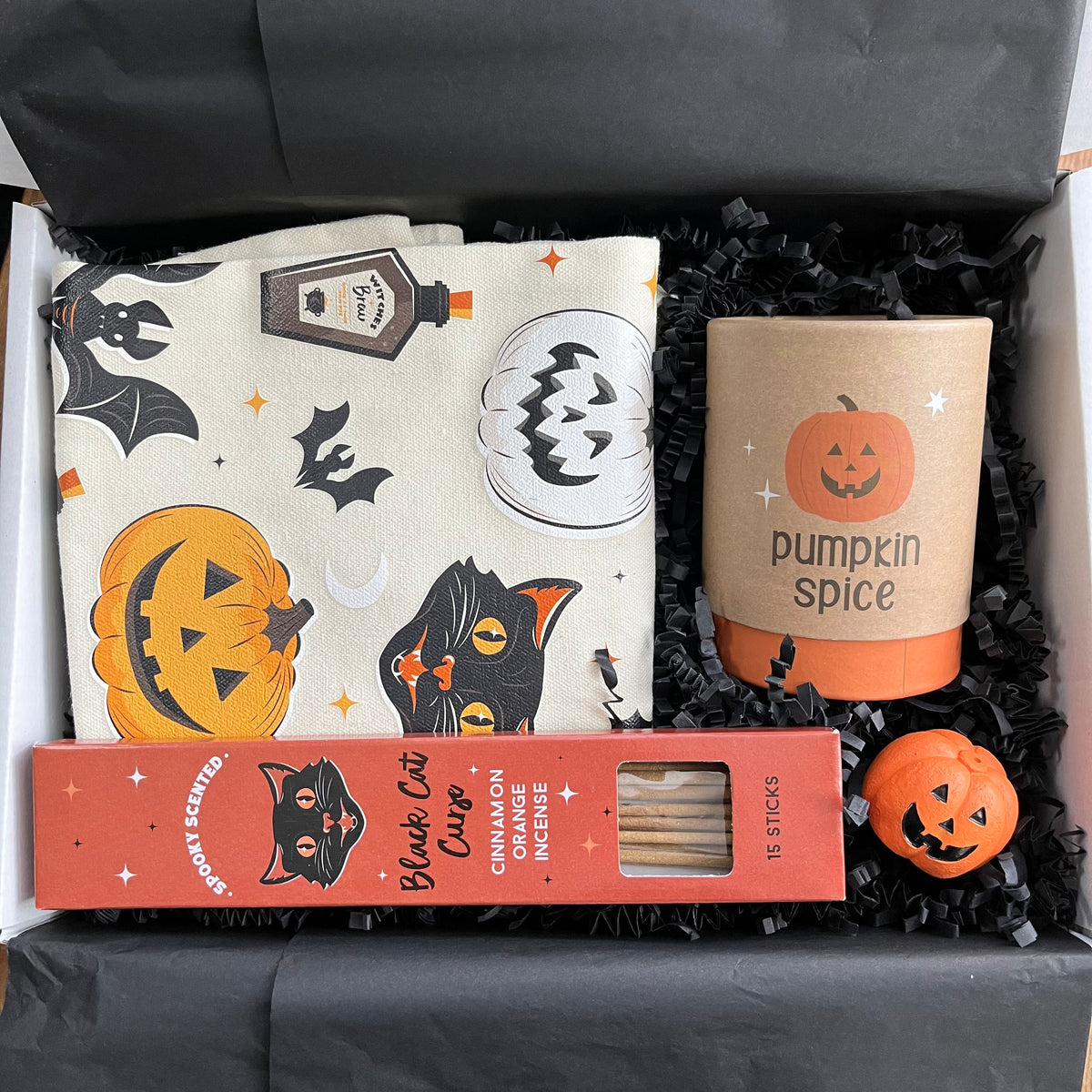 A curated Halloween gift box filled with black shredded paper, containing a cream fabric tote bag with a black cat and pumpkin print, a boxed candle labeled 'pumpkin spice' a red box of 'Black Cat Cinnamon Orange incense Sticks' , and a small orange jack-o'-lantern incense holder