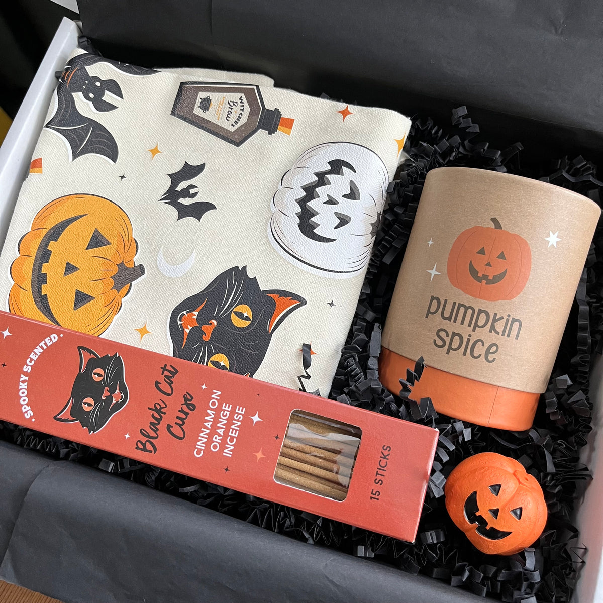 A curated Halloween-themed gift box containing a cream canvas tote bag with a black cat, jack-o'-lantern, and bat print; a boxed candle labeled 'pumpkin spice', box of 'Black Cat Cinnamon Orange' incense sticks; and a small orange jack-o'-lantern incense holder  all arranged in black shredded paper.