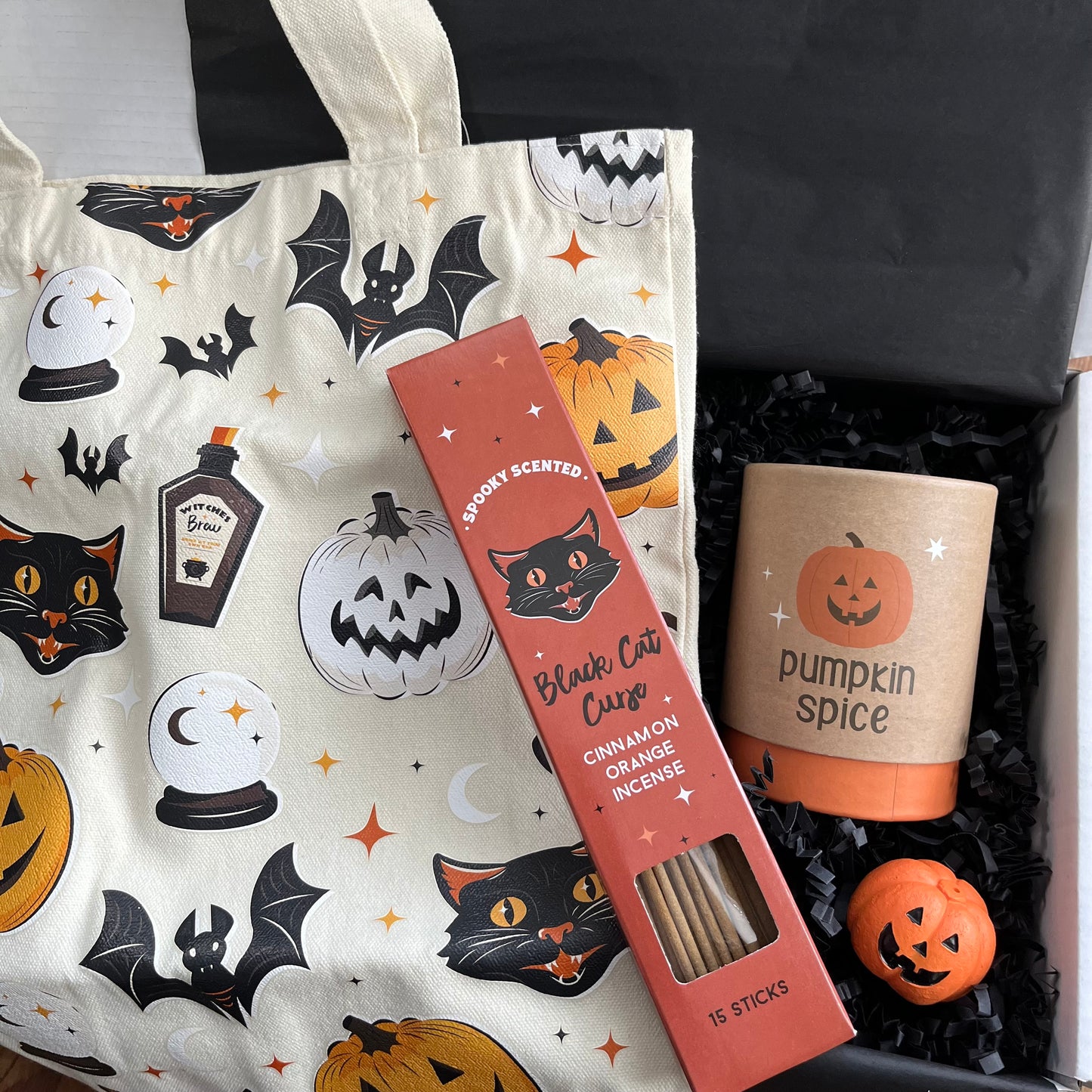 A Halloween-themed gift box filled with seasonal items, including a cream canvas tote bag with black bats, cats, pumpkins, a red box of 'Black Cat Curse' cinnamon orange incense sticks; a candle labeled 'pumpkin spice'; and a small orange jack-o'-lantern incense holder.