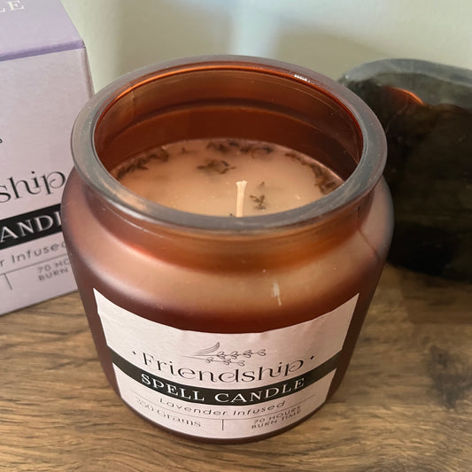Candle in a bronze jar labeled 'Friendship Spell Candle' on a wooden surface.
