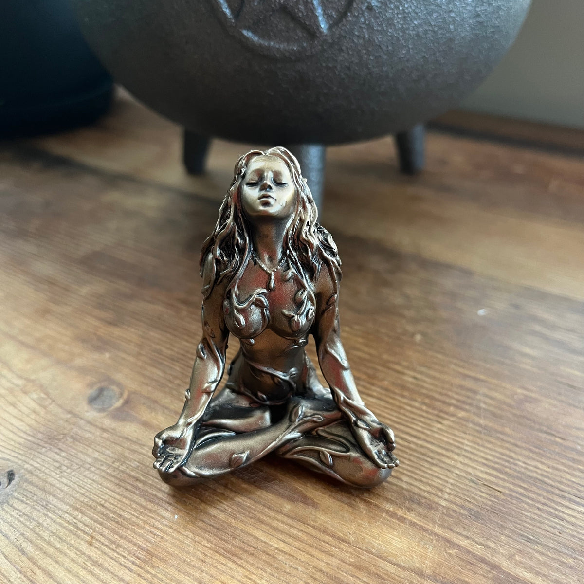 A small bronze figurine of a seated female figure with long flowing hair, displayed on a wooden surface with a dark cauldron in the blurred background.