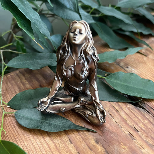 A bronze-finished figurine of a meditating female figure with long hair, styled on a wooden surface with green leaves.